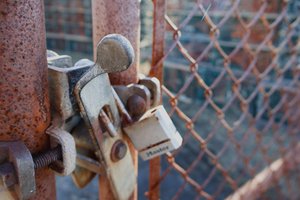 A close-up photo of a rusty metal gate secured with an old latch and a weathered white padlock. The background shows a blurred chain-link fence and industrial structures, slightly out of focus. The lighting highlights the textures of rust and metal wear.
