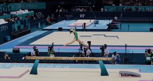 A gymnast in a green leotard leaps gracefully across a balance beam during the Paris 2024 Olympics. The arena is brightly lit with a blue and teal color scheme, and multiple photographers and cameramen are capturing the moment from below. The gymnast is midair with arms extended, and the stands are partially filled with spectators.