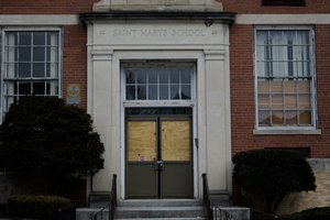 A photo of the boarded-up front entrance of an old brick school building labeled “Saint Mary’s School.” The door is covered with plywood, and the windows are cracked or obscured by blinds. A fallout shelter sign is mounted on the wall near the entrance. Overgrown bushes frame the doorway, adding to the abandoned atmosphere.