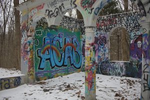 A photo of a snow-dusted, partially ruined concrete structure covered in colorful graffiti. The central wall features large blue letters reading “ACAB” with a rainbow painted over them. Surrounding walls and pillars are layered with various tags, messages, and images, including a white skull on a dark red background. Bare trees stand in the background, suggesting the scene is set in a quiet winter forest. The photo is unedited and retains natural lighting and color tones.